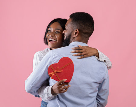 Excited black woman holding heart shaped gift box and hugging her boyfriend on pink studio background. Lovely African American couple celebrating Valentines Day togetherの写真素材