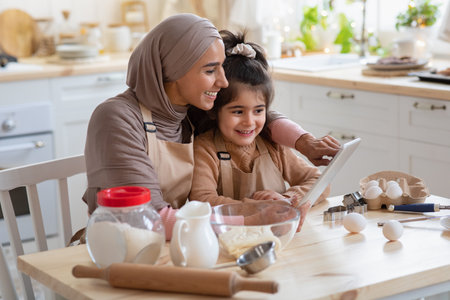 Smiling Muslim Mom And Her Little Daughter Using Digital Table In Kitchen, Checking Dough Recipe Online While Preparing Pastry Together, Happy Islamic Mother And Cute Girl Cooking At Home, Free Spaceの写真素材