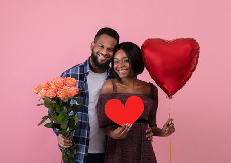 Valentines Day. Young black couple with red heart, balloon and bouquet of roses posing on pink studio background. Happy millennial woman with her boyfriend celebrating lovers holidayの写真素材