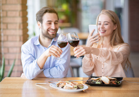 Portrait of lovely lady taking picture of toast with clinking wine glasses during holiday dinner at restaurant. Romantic millennial couple celebrating anniversary or Valentines Day, saying cheersの写真素材