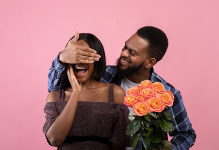 Loving black man surprising his girlfriend with bouquet of roses, covering her eyes over pink studio background. Affectionate African American couple with flowers celebrating Valentines Dayの写真素材