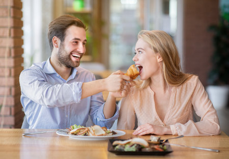 Affectionate young man feeding tasty croissant to his girlfriend at cozy cafe. Loving millennial couple having fun during dinner at coffee shop, spending happy times together, enjoying their mealの写真素材