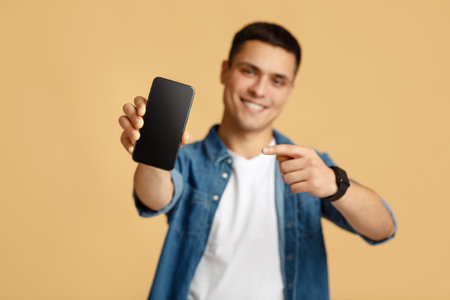 It is cool, best mobile app and digital ad. Happy young man in casual shows finger at smartphone with blank screen, selective focus on device, isolated on beige background, studio shot, free spaceの写真素材