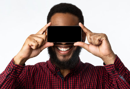 Look At This App. Black Guy With Smartphone Posing, Holding Phone Screen In Front Of Face Standing On White Studio Background. Man Covering Eyes With Cellphone Advertising Mobile Application. Mockupの写真素材