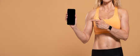 Fitness advice from popular coach blogger and mobile workout app. Muscular middle-aged female in sports uniform and fitness tracker shows smartphone with blank screen, isolated on beige backgroundの写真素材