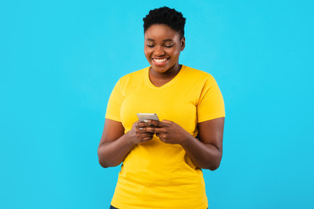 Smiling Black Oversized Lady Using Smartphone Texting Standing Over Blue Studio Background. African...