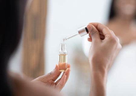 Perfect skin, natural cosmetic product for hydrated, glowing and healthy facial derma. African american lady with applying essential oil with dropper on face and look at mirror, blurred, croppedの写真素材