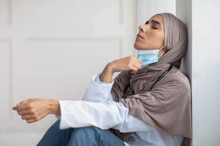 Tired muslim woman doctor removing protective face mask, feeling exhausted after difficult working day, sitting by wall with closed eyes at clinic, copy space. Doctors and COVID-19 pandemic conceptの写真素材