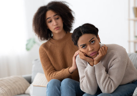 Relationship problems, quarrel and forgiveness at self-isolation. Caring millennial african american lady calm and support offended and angry girlfriend, sitting on sofa in living room interiorの写真素材