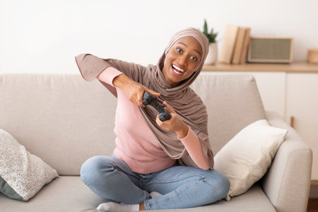 Fun stay home pastimes. Happy black woman in hijab using joystick to play online video games on...