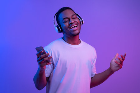 Joyful black guy in wireless headphones listening music online on smartphone and singing under futuristic neon lighting, enjoying favorite songs, standing with closed eyes on fluorescent backgroundの写真素材