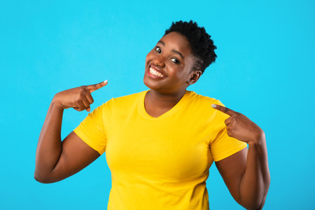 Self-Confidence. Happy Plus-Size Black Woman Pointing Fingers At Herself Posing Standing Over Blue Studio Background, Smiling To Camera. Choose Me, Im The Best Conceptの写真素材