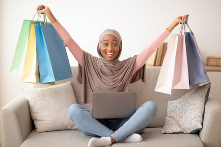 Cheerful black lady in hijab shopping on internet from laptop, lifting paper bags up, feeling excited at home. Happy Muslim woman in headscarf buying goods in web store, ordering onlineの写真素材