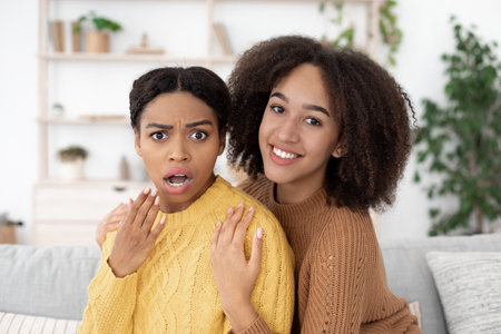 Gossip, shocking news, unusual ad and pain at home. Smiling millennial african american lady hugs surprised girlfriend and look at camera, sitting on sofa in living room interior, close up, free spaceの写真素材