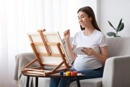 Painting While Pregnant. Happy Young Expecting Woman Drawing Picture On Easel At Home, Enjoying Art Therapy And Creative Leisure During Pregnancy, Young Awaiting Lady Sitting On Couch In Living Roomの写真素材