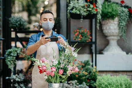 Flower shop, delivery, creating order and modern online sale. Millennial busy male owner in apron and protective mask makes fashionable bouquet and takes photo, in studio, empty space, outdoorの写真素材