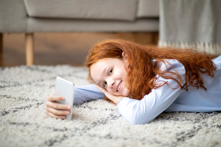 Homeland Generation. Portrait of happy smiling young teenage girl holding and using smartphone while lying on the floor carpet in bedroom, resting and relaxing at home, looking at camera, chattingの写真素材