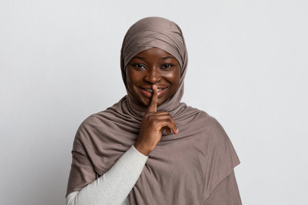Mysterious Black Muslim Woman In Hijab Showing Shh Sign, Putting Finger On Lips, Smiling African Religious Lady In Headscarf Gesturing Hush And Keep Silence Over Light Studio Background, Free Spaceの写真素材