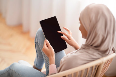 Black Arab woman in hijab using tablet computer with empty screen while sitting in cozy armchair at home, mockup for website design. Muslim lady with touch pad working or studying onlineの写真素材