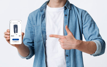 Mobile Shopping App For Customers. Closeup of casual young man holding and pointing at smartphone with clothes shop website, ordering via internet.の写真素材