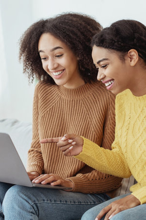 Amazing event, new interesting message, ad and great online offer. Cheerful millennial african american girlfriends points finger at laptop, sits on couch in living room, vertical, close upの写真素材