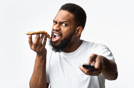Hungry African American Man Eating Pizza And Watching TV Pointing Television Remote Control To Camera Posing On White Studio Background. Junk Food, Unhealthy Lifestyle, Overeating And Nutrition Habitsの写真素材