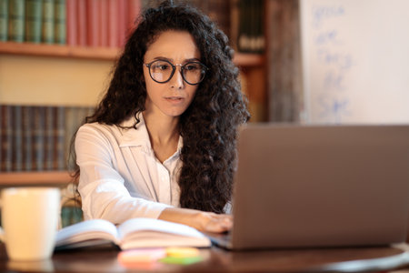 Portrait of pensive and confused woman in eyeglasses using and working on computer, typing on laptop keyboard. Puzzled young lady sitting at desk, doing work project and thinking about deadlineの写真素材