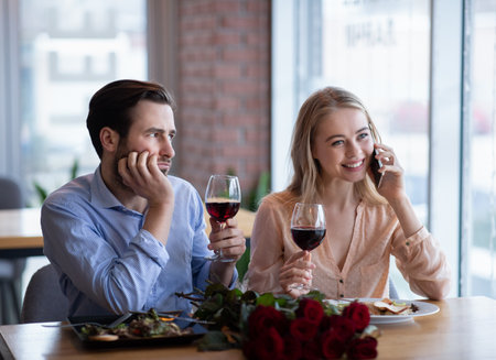 Millennial lady talking on cellphone during boring date, young guy feeling neglected and upset at restaurant. Couple of lovers having problems during romantic Valentines dinner with roses and wineの写真素材
