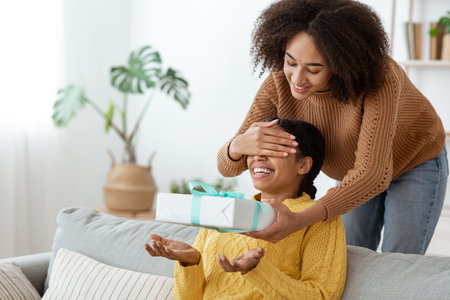 Birthday with friend during covid-19 quarantine. Cheerful mixed race lady, makes surprise, closes eyes and gives box with ribbon to excited african american woman in modern living room interiorの写真素材