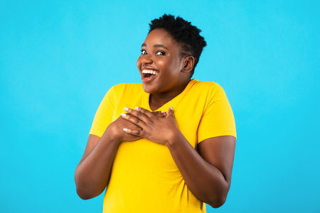 Excited Oversized African American Lady Posing Holding Hands On Chest Smiling Looking At Camera In Excitement Standing Over Blue Studio Background. Pleasant Surprise, Positive Emotions Conceptの写真素材