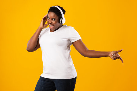 Positive Overweight Black Lady Listening To Music In Wireless Headphones Enjoying Favorite Song Posing Standing Over Yellow Studio Background, Smiling To Camera. Great Playlist Conceptの写真素材