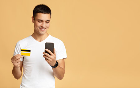 Large sale, modern online shopping and buying. Cheerful millennial male student in white t-shirt holds credit card and pays with smartphone, isolated on beige background, studio shot, free spaceの写真素材