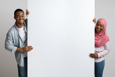 Cheerful african american muslim couple posing with big empty board over grey studio background. Smiling young black man and woman in pink hijab standing next to blank advertising placardの写真素材