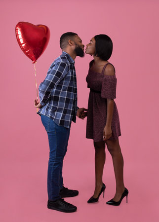 Full length portrait of loving black couple kissing, celebrating Valentines Day together on pink studio background. African American guy with heart shaped balloon being affectionate to his girlfriendの写真素材