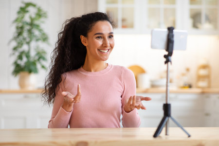 Blogging Concept. Female Blogger Using Smartphone On Tripod For Recording Video At Home, Young Cheerful Woman Sitting In Kitchen Interior, Talking And Gesturing At Camera, Smiling Lady Capturing Vlogの写真素材