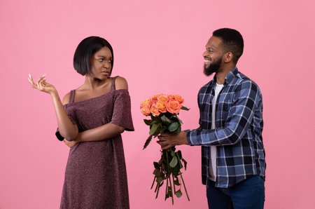 Irritated black woman rejecting geek man offering flowers on Valentines Day over pink studio background. Black guy asking for forgiveness after fight with bouquet of roses, young lady refusing himの写真素材