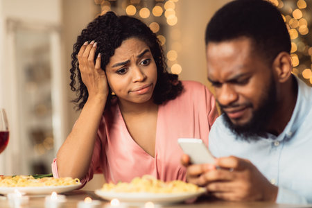 Bad Date. Unhappy Sad African American Woman Is Getting Bored Sitting On Date In Restaurant While Her Boyfriend Using Mobile Phone And Chatting, Ignoring His Girlfriend. Relationship Problemの写真素材