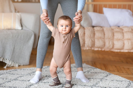Cute little baby boy or girl taking first steps with mother help, home interior, close up. Adorable...