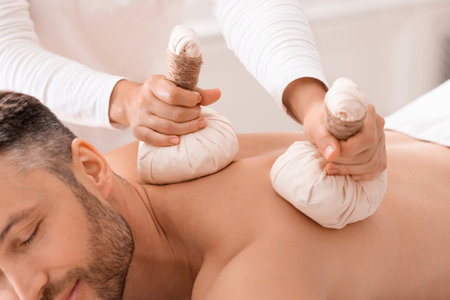 Closeup of compress balls in unrecognizable masseuse hands making herbal massage for handsome bearded man. Middle-aged man spending day at male spa, getting healing herbal massageの写真素材