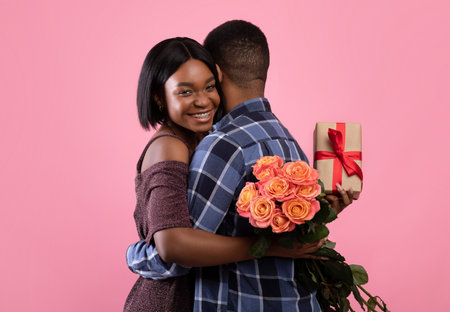 Passionate African American lovers with gift box and beautiful roses hugging on pink studio background. Affectionate black couple celebrating Valentines Day or anniversary togetherの写真素材