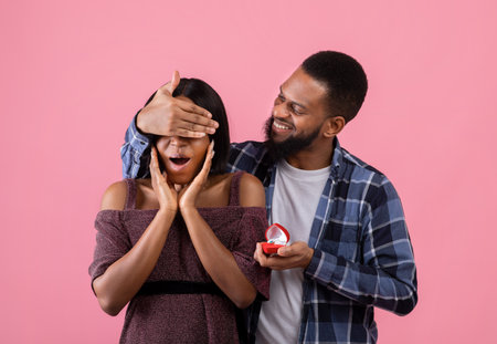 Affectionate black guy covering his sweethearts eyes, giving her engagement ring, making marriage...