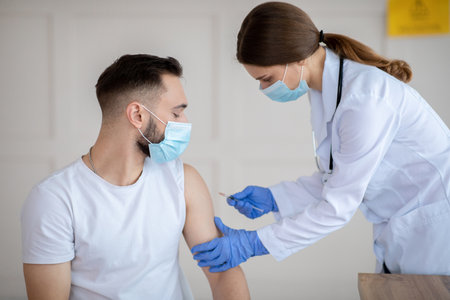 Vaccination against covid-19. Female doctor in face mask injecting coronavirus vaccine to male patients arm at health centre. Young man being immunized against global virus at clinicの写真素材