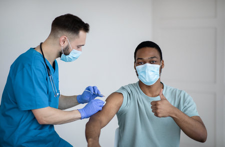 Black guy in face mask gesturing thumb up during covid-19 vaccination, approving of coronavirus...