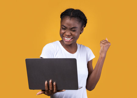 Joyful African American Lady Holding Laptop Gesturing Yes Celebrating Success And Great News Standing Posing Over Yellow Studio Background. Joy, Gadgets Lifestyle Conceptの写真素材