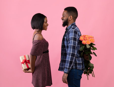 Side view of positive black couple with flowers and gift for Valentines Day looking at each other over pink studio background. Passionate sweethearts hiding holiday surprises behind their backsの写真素材