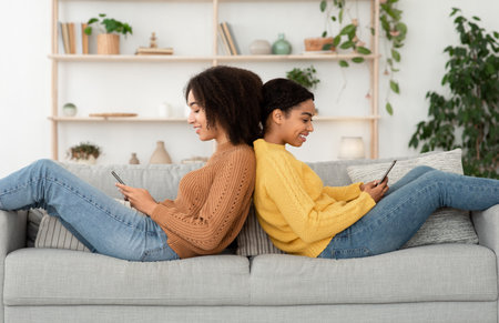 Free time, browsing and surfing in internet together. Smiling cute young african american women sitting back to back on sofa and typing messages on smartphones in living room interior, side viewの写真素材
