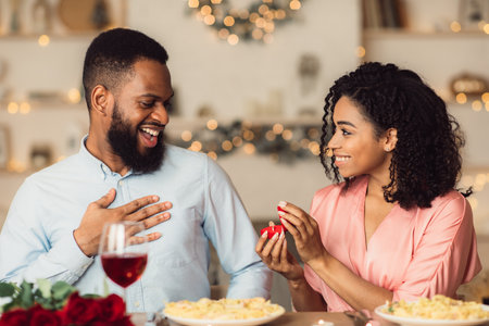 Will You Marry Me. Smiling african american woman proposing to her boyfriend, holding and showing open box, offering marriage ring to happy man, getting engaged in restaurant during romantic dateの写真素材