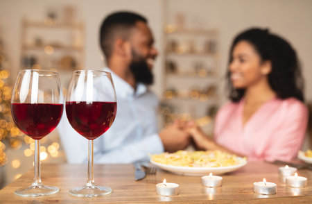 Romantic Date. Selective focus on two glasses of red wine on the table in the foreground. Black couple spending time together at home or cafe, having dinner, holding hands in blurred backgroundの写真素材