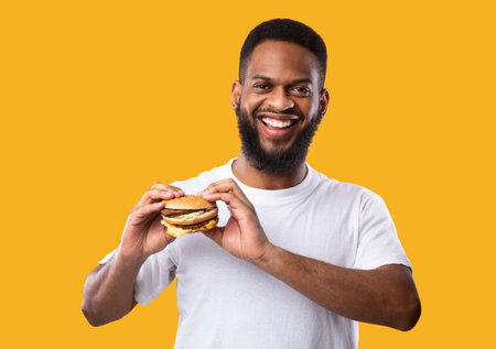 Happy black man posing holding burger smiling to camera standing over yellow background, studio shot. Junk food and cheat meal, nutrition and male diet concept. Overeating habitの写真素材