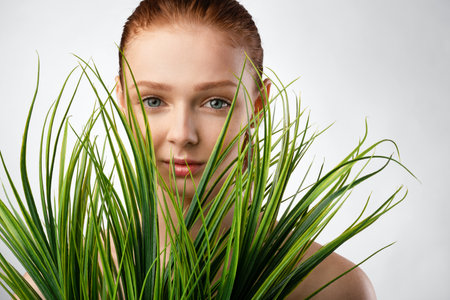 Natural Beauty. Red-Haired Girl Posing With Green Plants Looking At Camera Standing Over White Studio Background. Female Portrait. Youth, Wellness And Spa, Skin And Body Care Conceptの写真素材